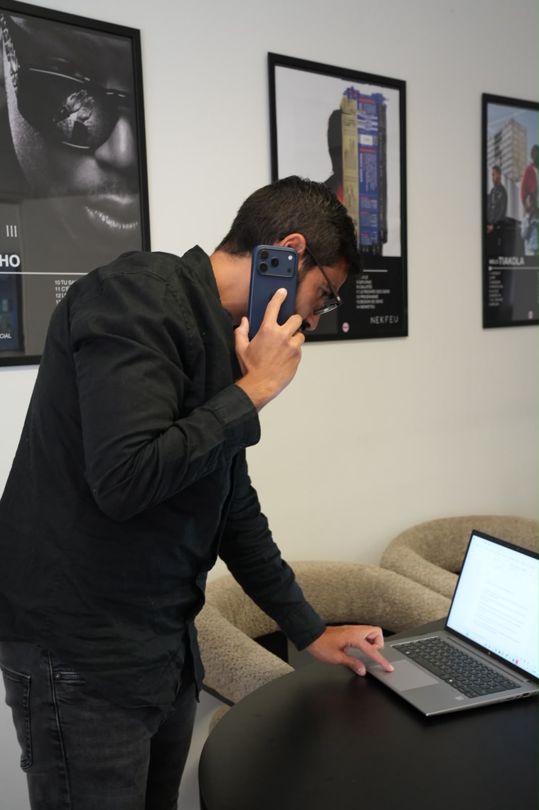 Homme en chemise noire parlant au téléphone tout en utilisant un ordinateur portable sur une table noire.