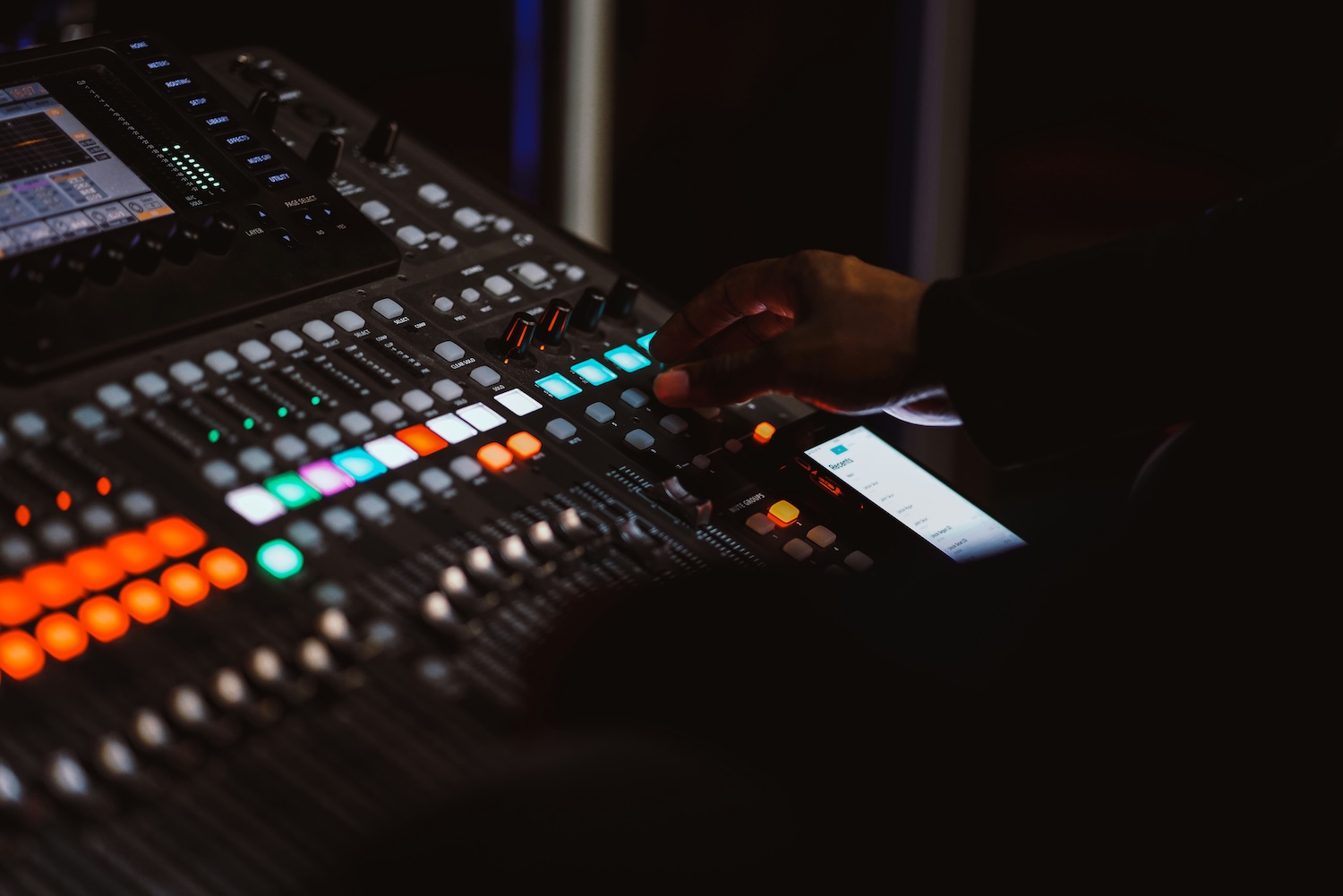 Main d'une personne ajustant des boutons lumineux sur une table de mixage audio dans une pièce sombre.