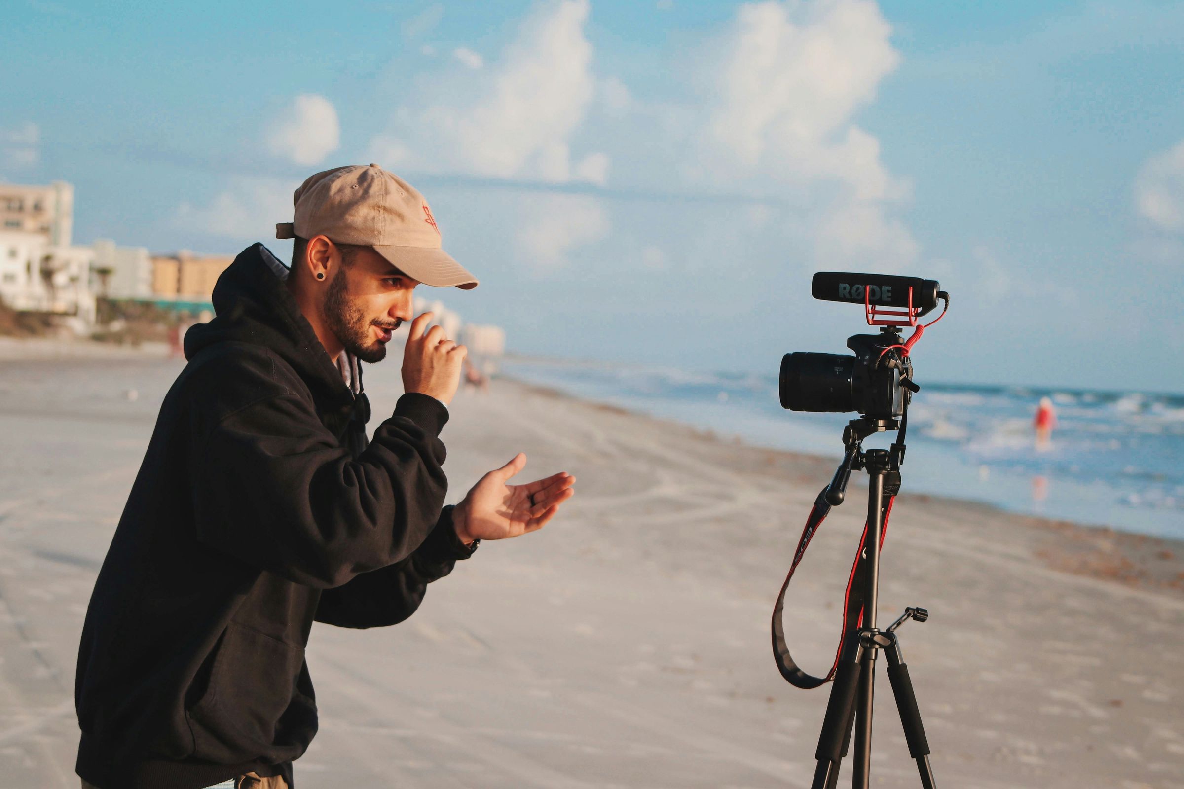 Un homme en sweat noir et casquette filme avec un appareil sur un trépied sur une plage.