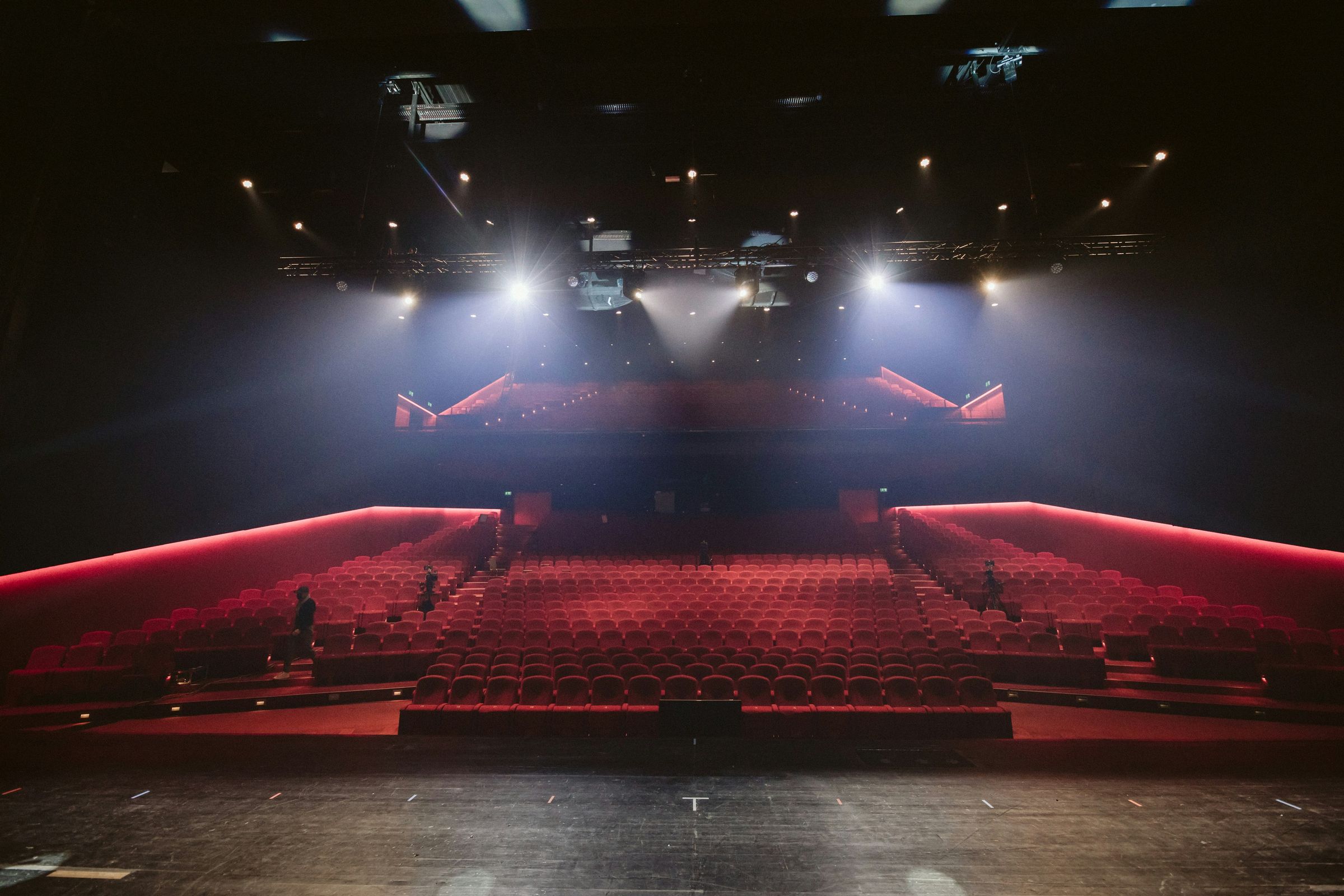 Salle de théâtre vide avec sièges rouges et des projecteurs allumés sur la scène.