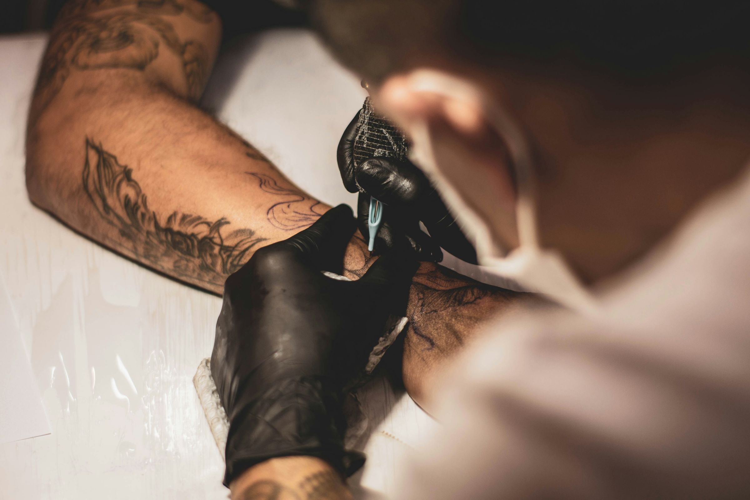 Un tatoueur portant des gants noirs tatoue l'avant-bras d'un client allongé.