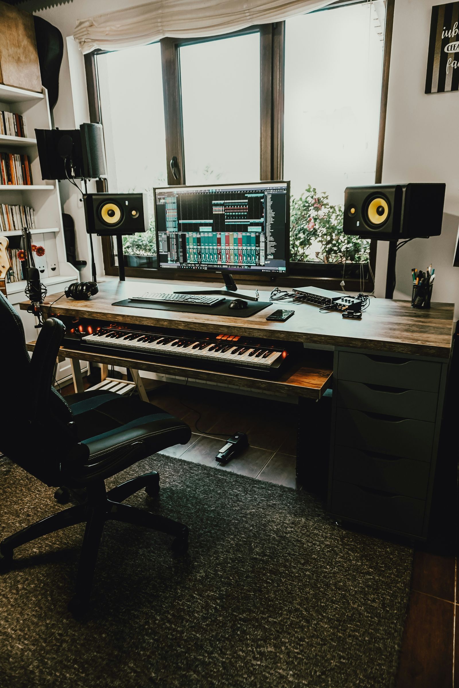 Bureau de production musicale avec clavier, moniteur, enceintes et microphone près de la fenêtre.