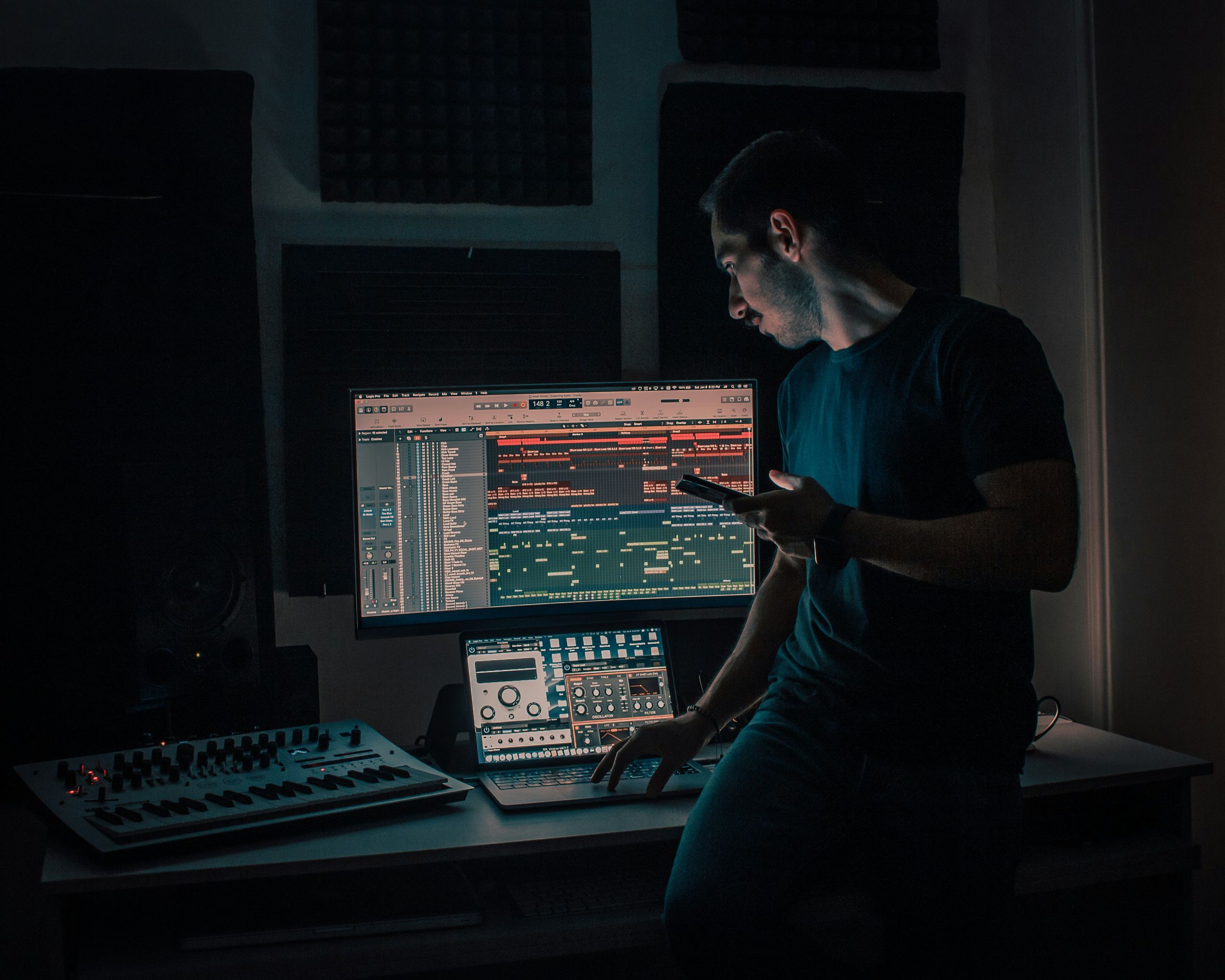 Homme utilisant un téléphone et un ordinateur avec logiciel de production musicale dans un studio sombre.