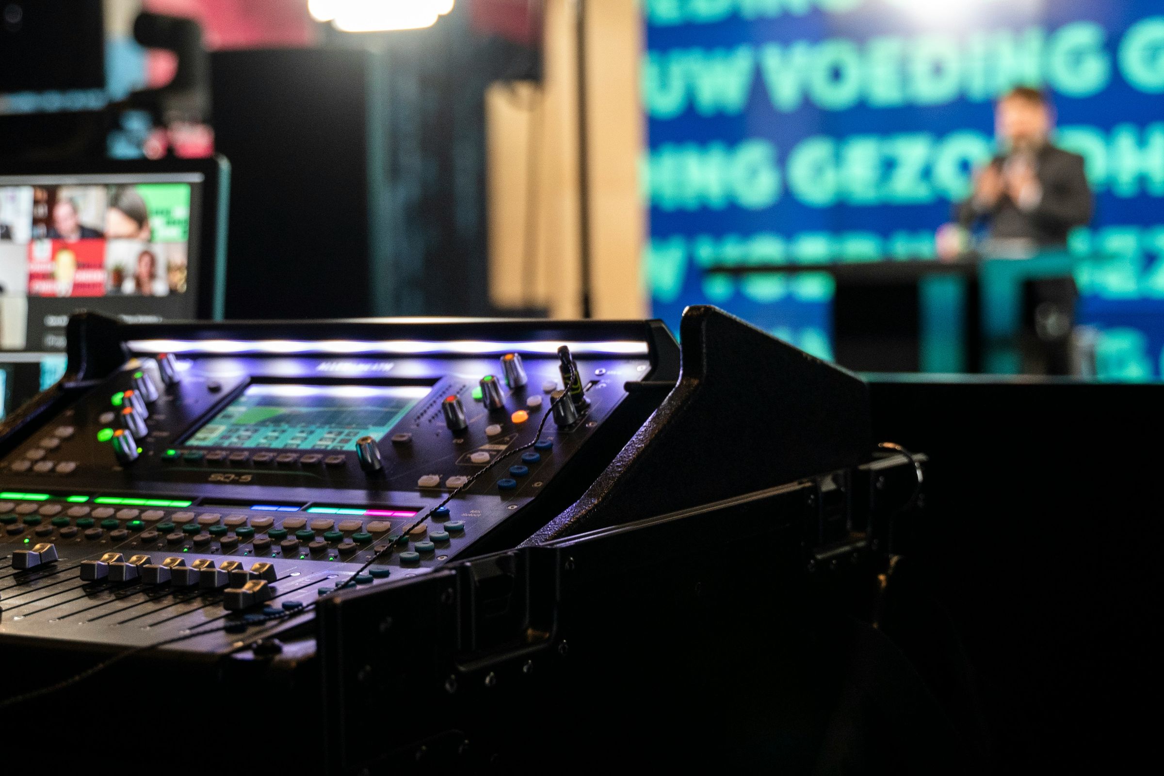 Console de mixage audio en gros plan avec un écran lumineux, un orateur flou en arrière-plan.