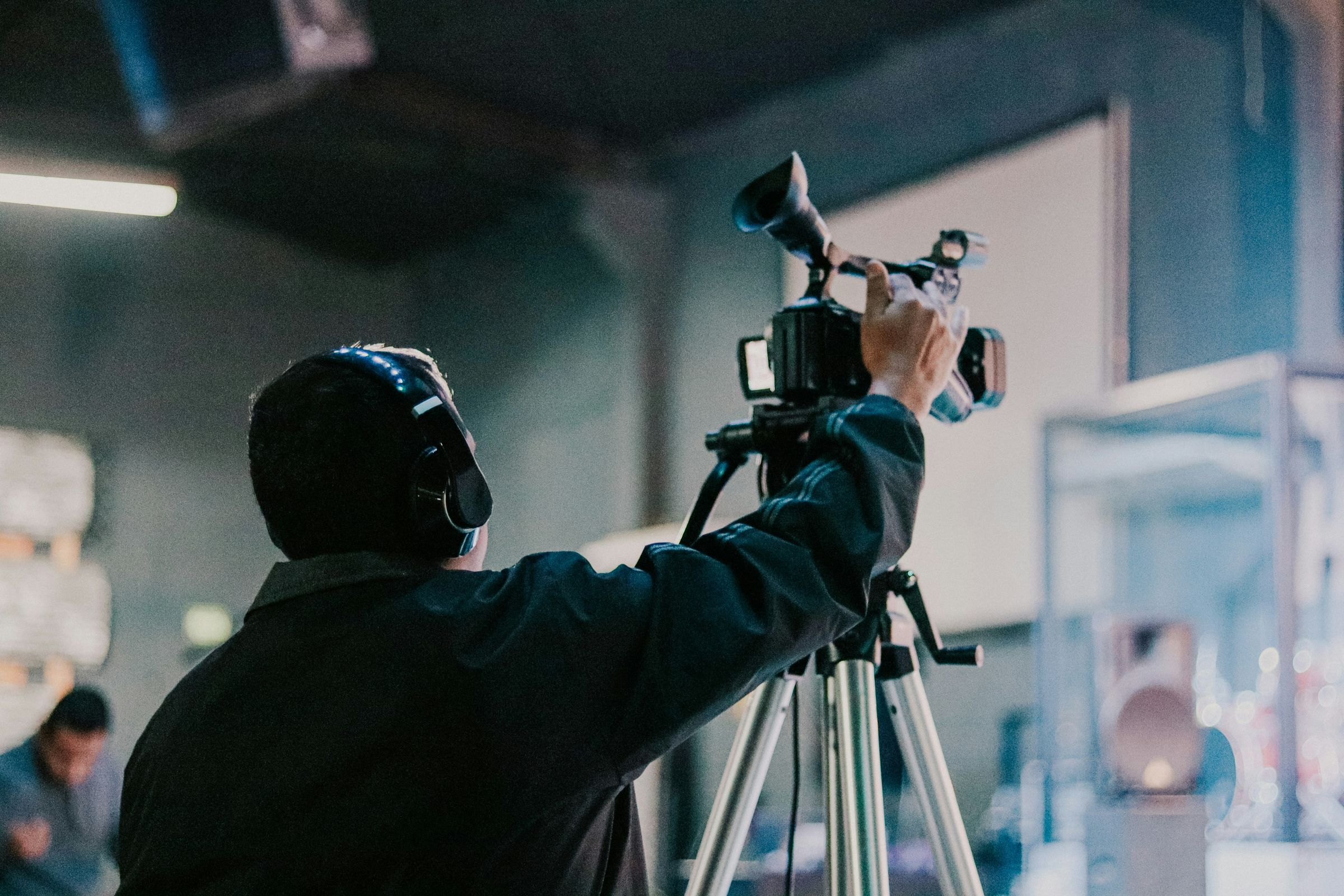 Cameraman adjusting a video camera mounted on a tripod while wearing headphones indoors.