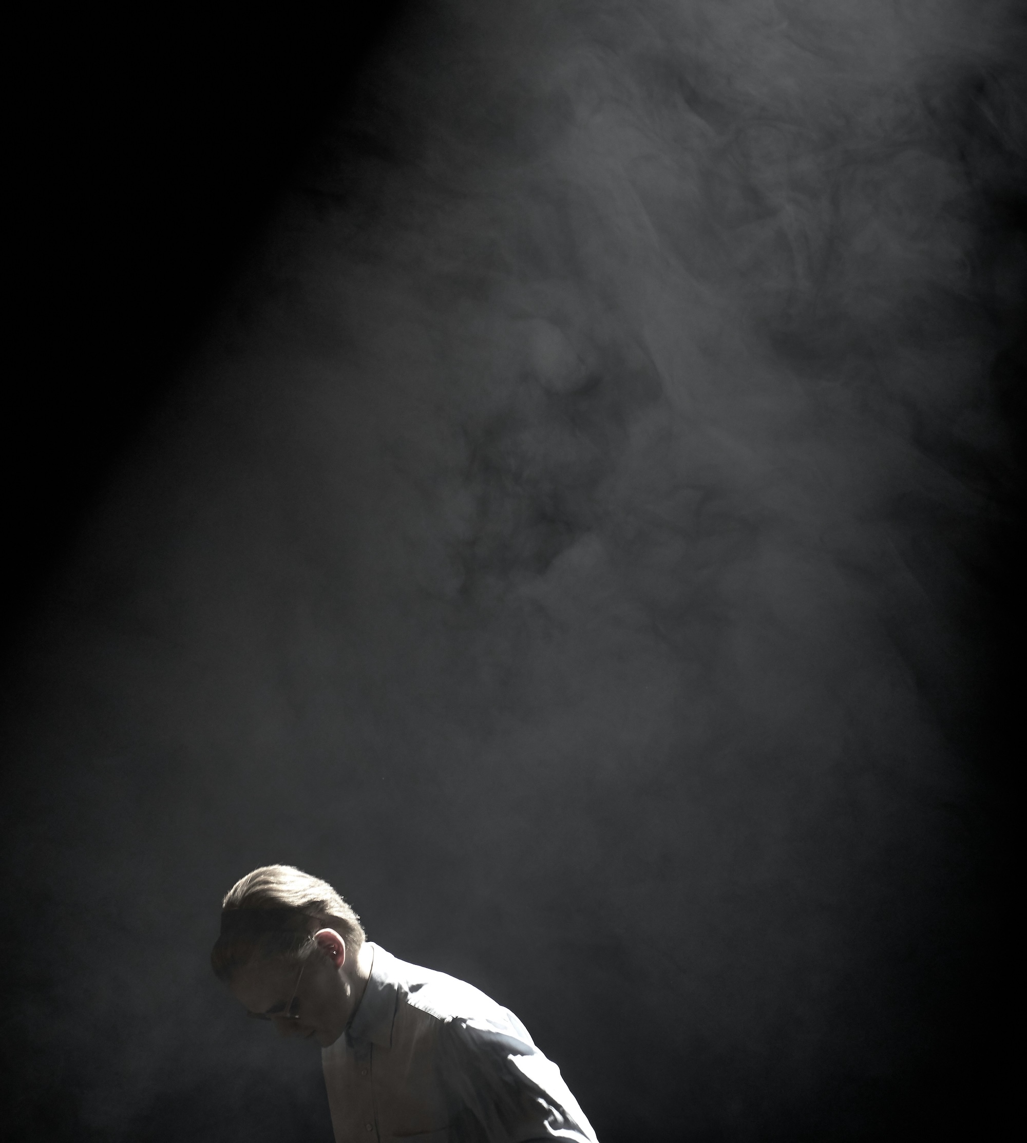 Homme penché en chemise blanche sous un faisceau de lumière dans une atmosphère sombre et enfumée.