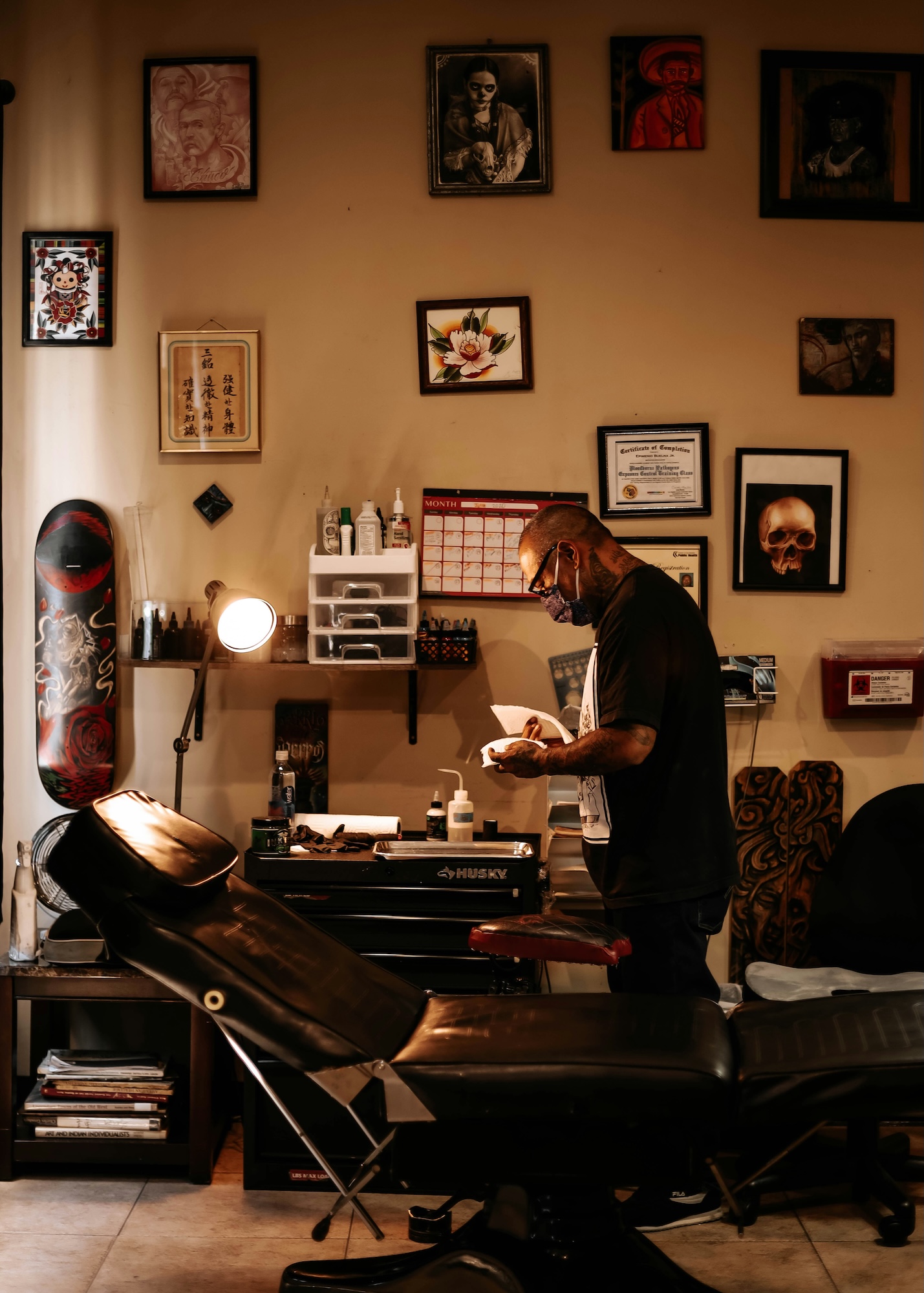 Tattoo artist wearing mask preparing tools by chair in a tattoo studio with framed art on the wall.