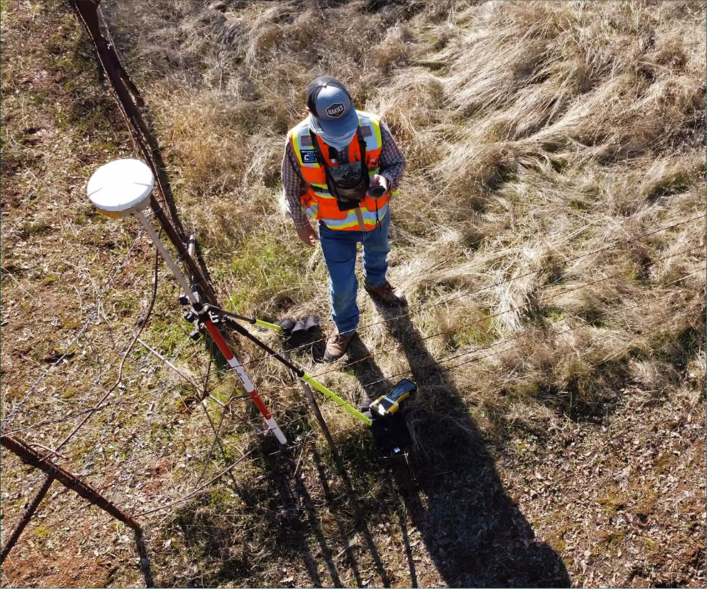 Surveyor wearing safety vest and cap using surveying equipment in grassy field near a fence.