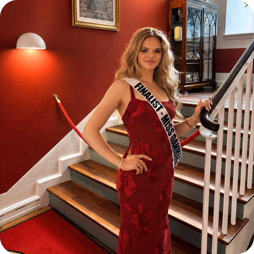 Young woman in a red gown with a finalist sash posing on stairs next to a red wall and display cabinet.