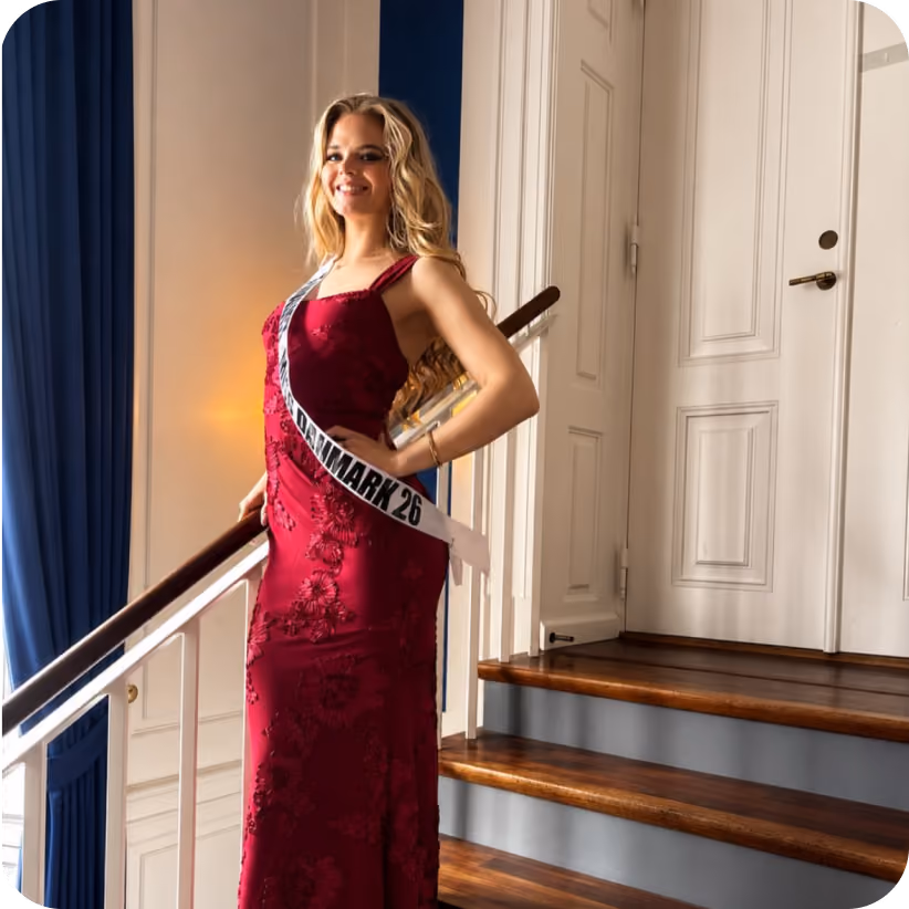 Woman in a red dress and sash reading Denmark 26 posed on wooden stairs inside a building.