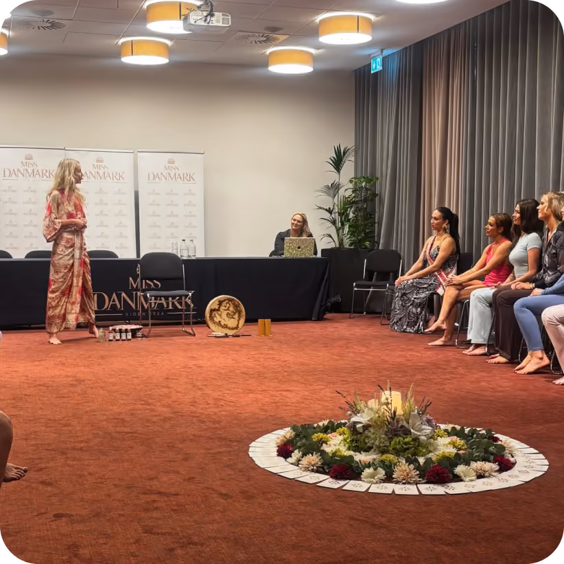 Women seated and listening to a barefoot woman standing and speaking in a carpeted room with Miss Danmark banners.