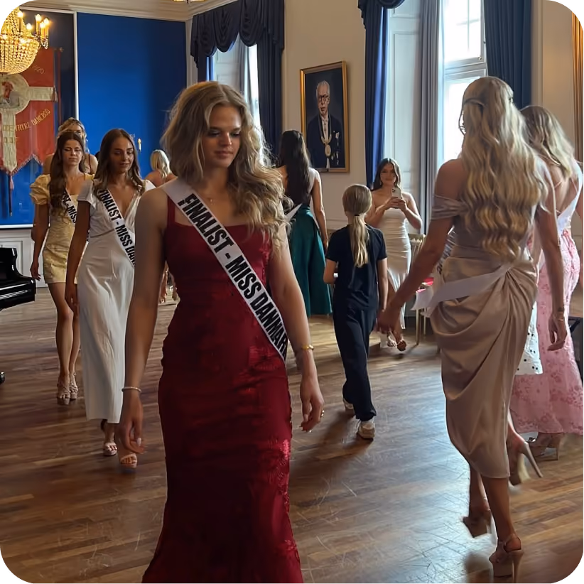 Women in elegant dresses wearing Miss Danmark sashes walking in a room with wooden floor and chandelier.