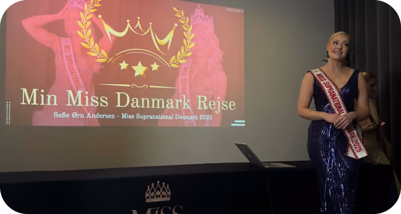 Woman in evening gown and sash stands beside a screen showing ‘Min Miss Danmark Rejse’ presentation.