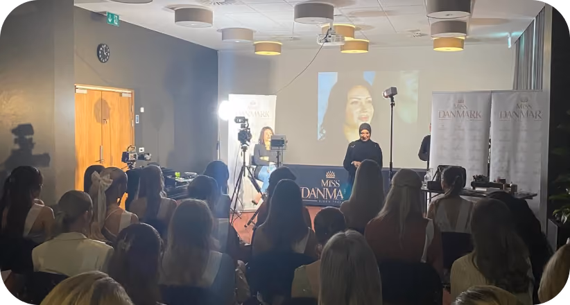 Audience watching a presenter at Miss Danmark event with camera and projection screen in a conference room.