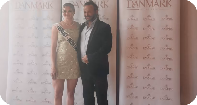 Woman in gold dress with finalist sash and man in dark suit posing in front of Danmark banners.