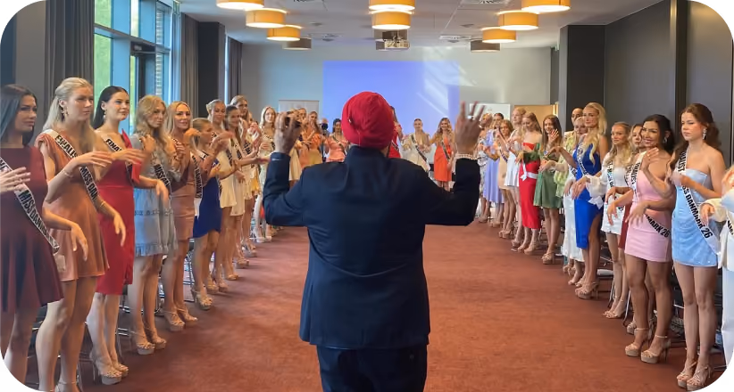 A group of women wearing sashes stand in two lines facing a person in a red headwrap and dark suit.