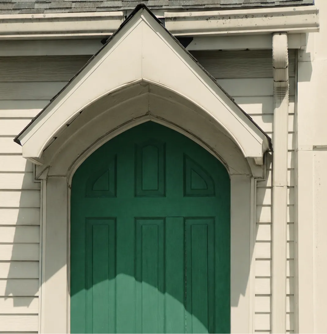 Green arched door set in light-colored siding with a white pointed overhang and shadows.