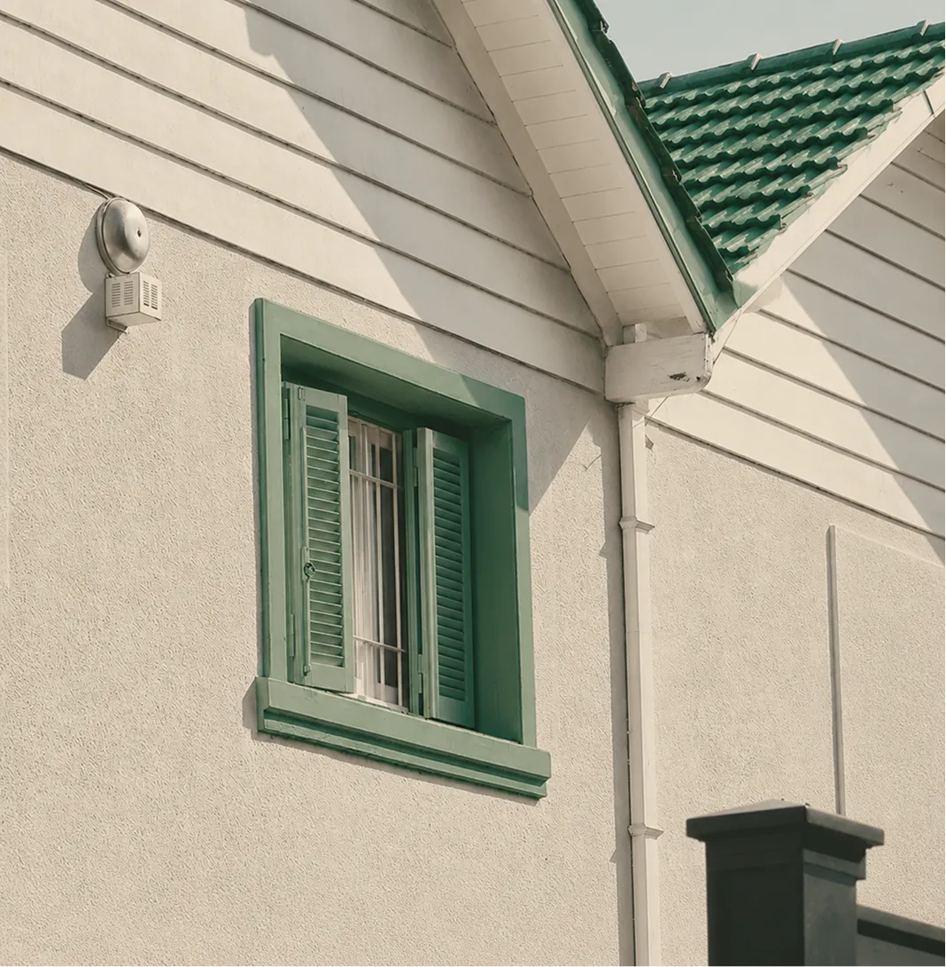 Beige house wall with a green window frame and shutters, beneath a green tiled roof.