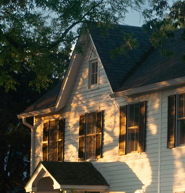 White house with black shutters in late sunlight filtering through tree branches.