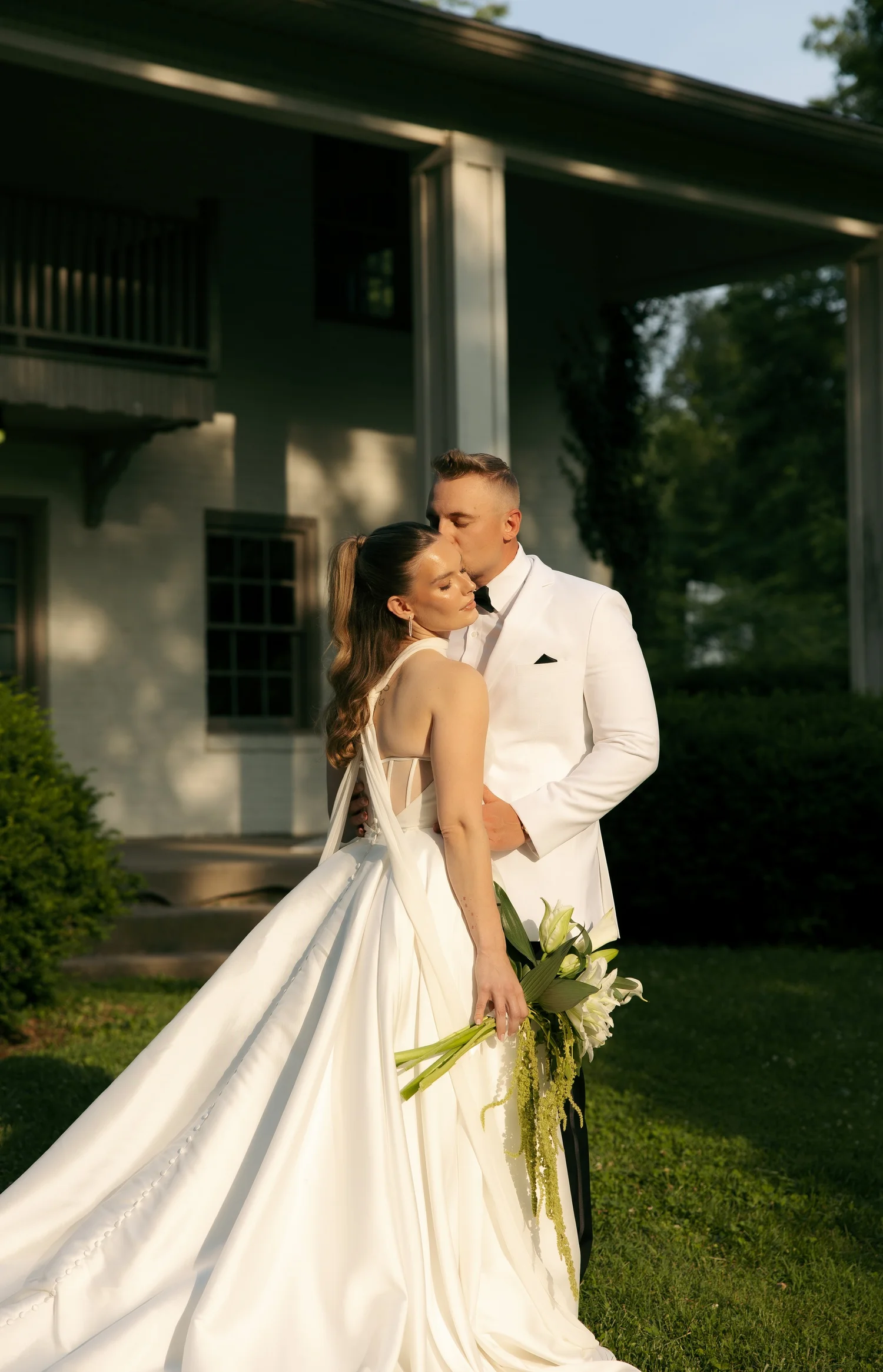 Man in white tuxedo kissing woman in white wedding gown holding lilies outdoors.