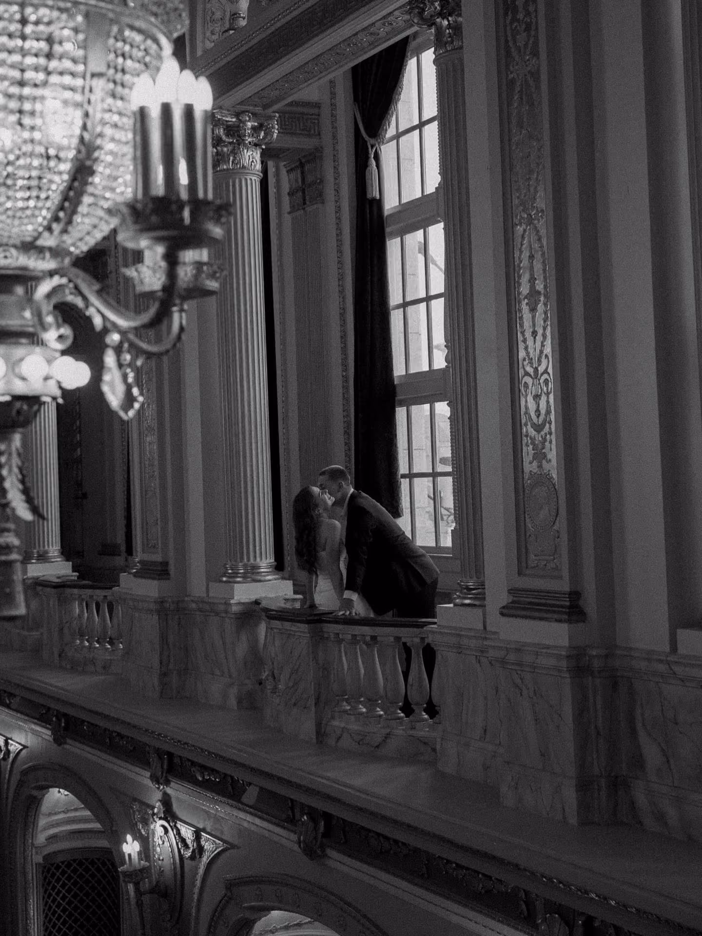 A couple sharing a romantic kiss on a marble balcony inside a grand, ornate building.