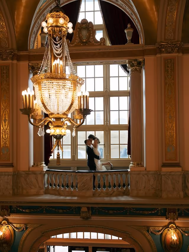 Couple kissing in elegant room with large windows and ornate golden chandelier above.
