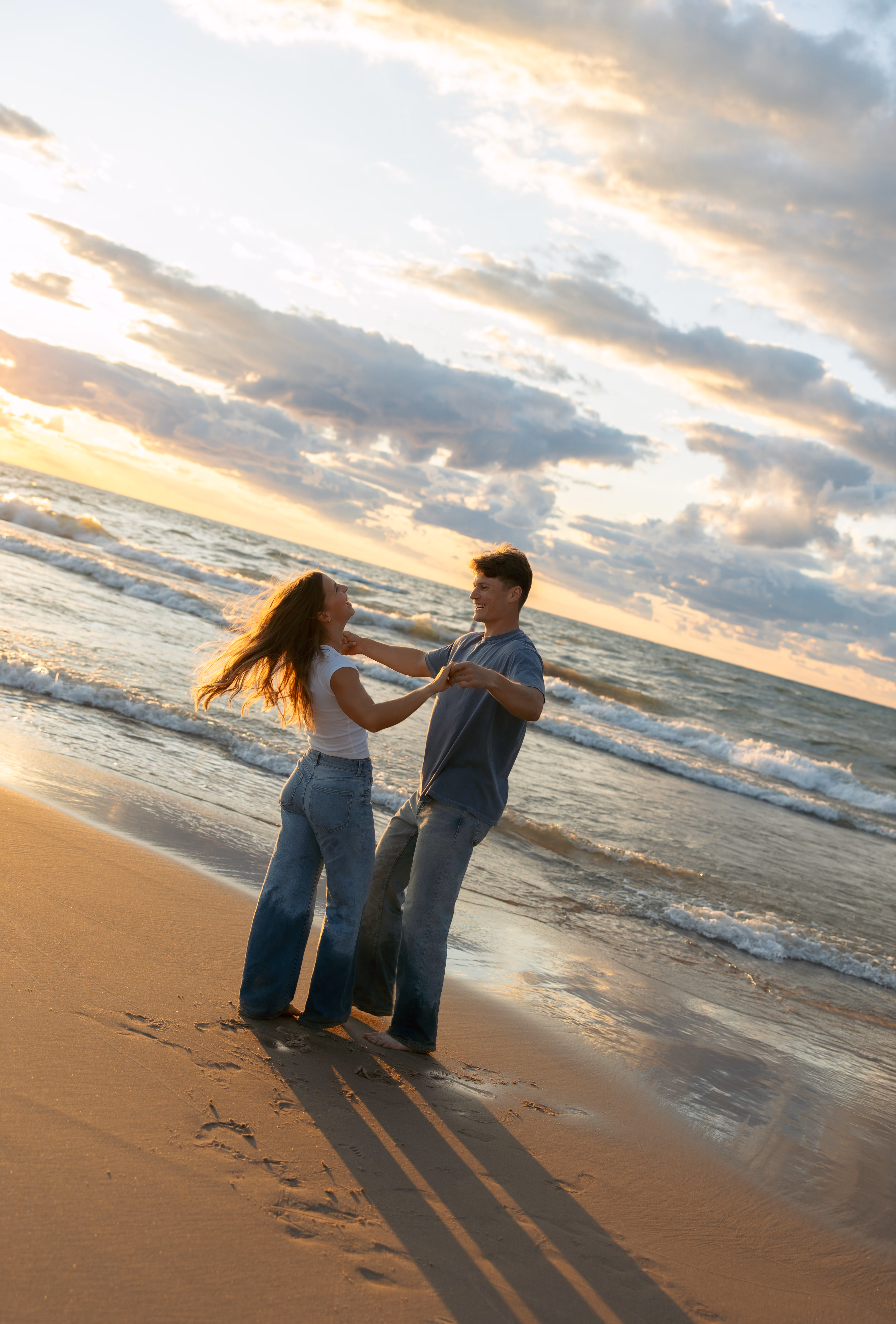 Couple holding hands and smiling while standing barefoot on a sandy beach at sunset.