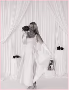 Woman in white dress holding a bouquet, standing against gathered white draped fabric backdrop.