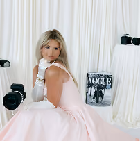 Woman in a light pink gown and white gloves poses among vintage cameras and an old Vogue magazine.