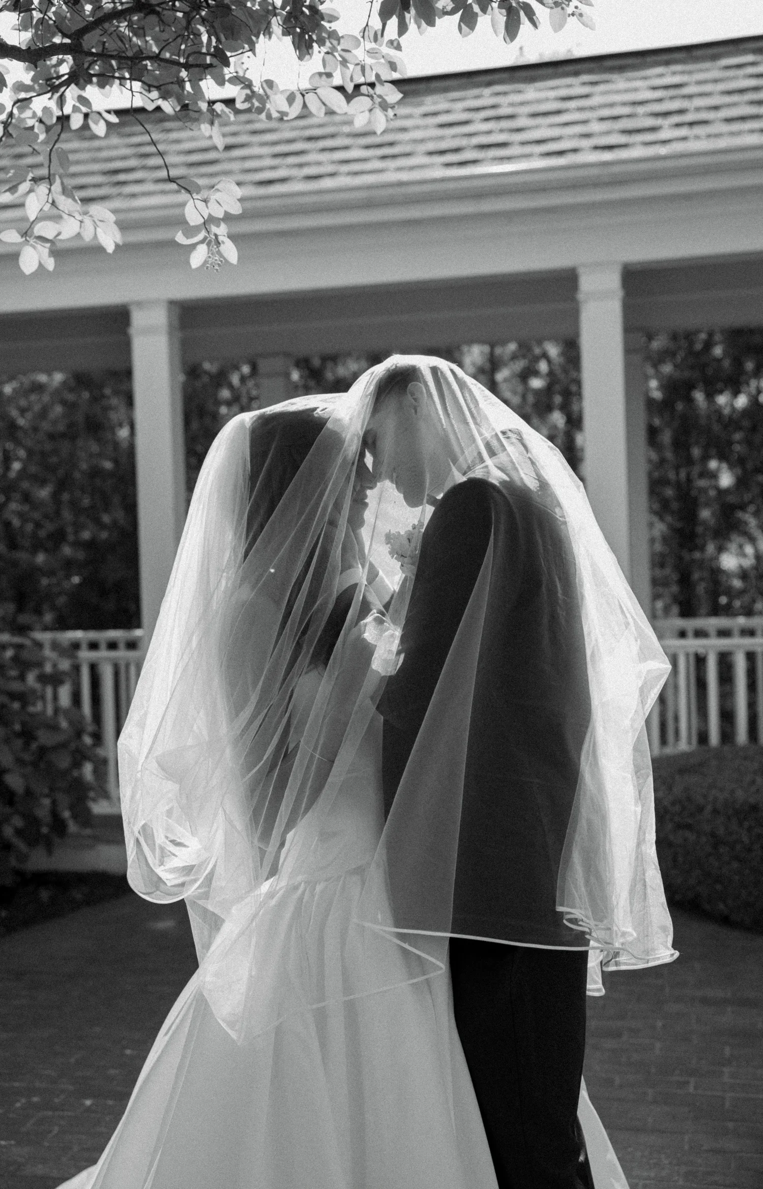Black and white photo of bride and groom under wedding veil, about to kiss outdoors.