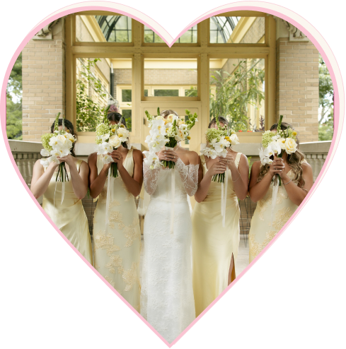 Bride in white dress and bridesmaids in cream dresses holding white flower bouquets in front of their faces.