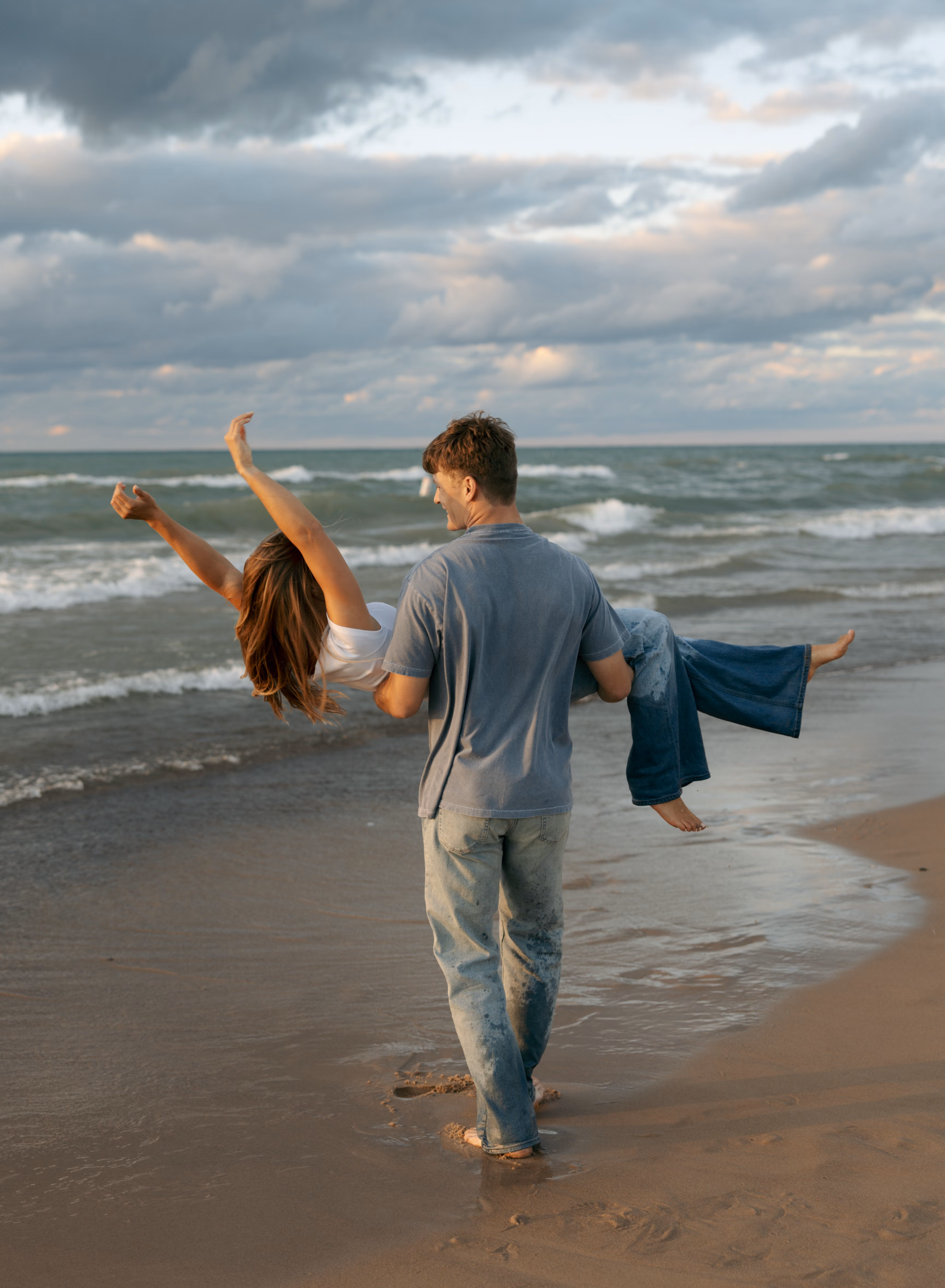 Man carrying woman with arms raised walking barefoot on sandy beach near ocean waves.