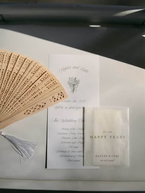 Wedding invitation with a wooden fan and a packet labeled 'for your happy tears' on a white surface.