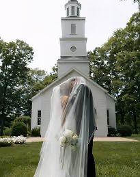 Bride and groom kissing under veil in front of white church with steeple and green trees.