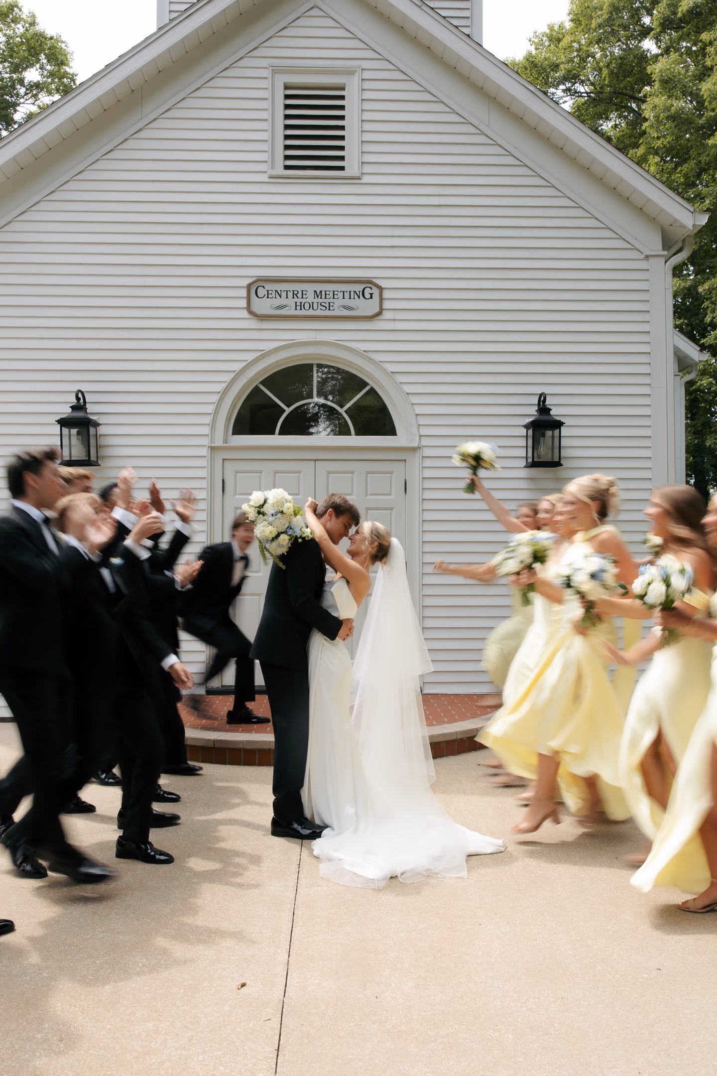 Bride and groom embrace while bridesmaids and groomsmen cheer outside white building.