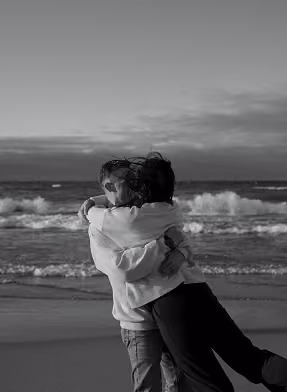Black and white photo of two people hugging on a beach with waves in the background.