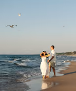 Couple walking on sandy beach near ocean waves under clear sky with flying birds.