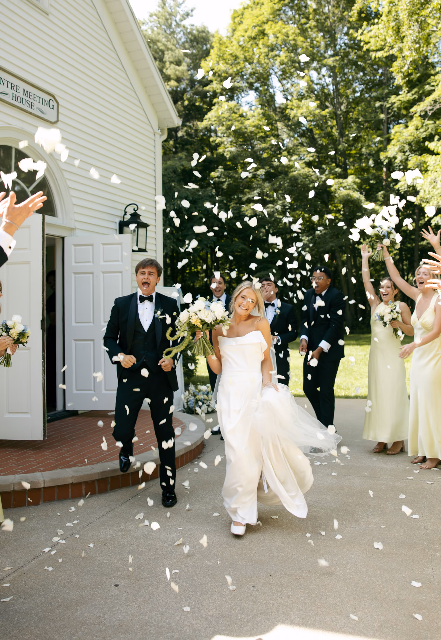 Bride and groom joyfully exit a building while guests throw white flower petals outdoors.