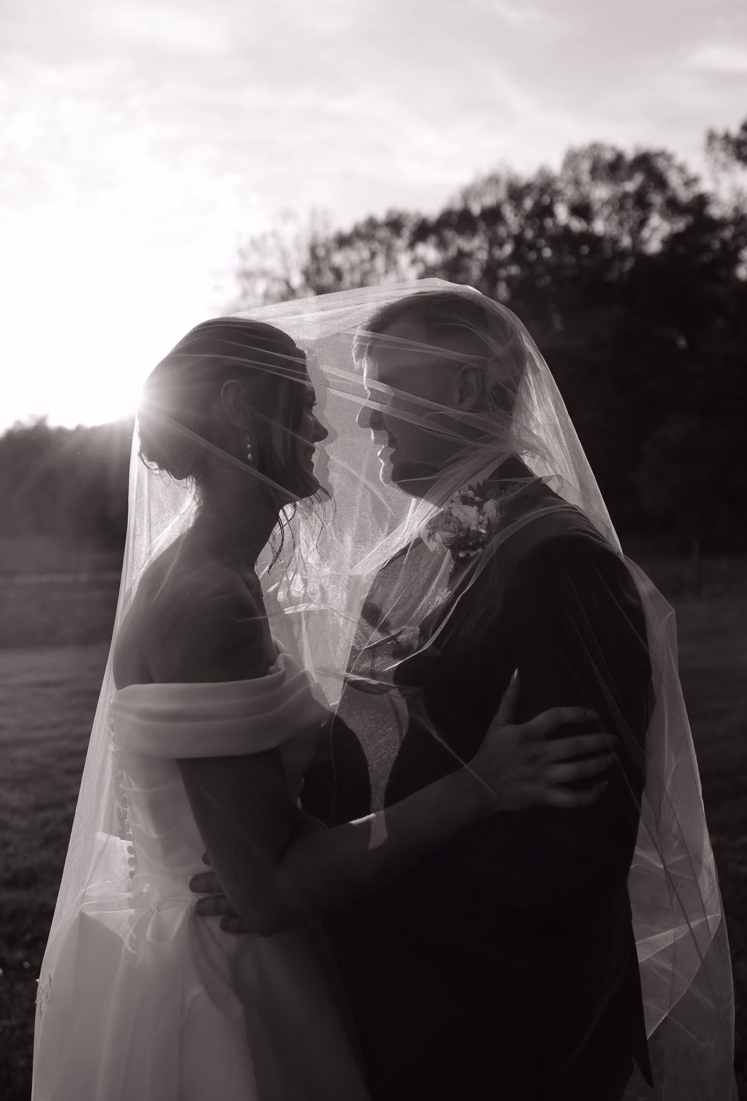 Bride and groom embracing face to face under veil with sunlight and trees in background.