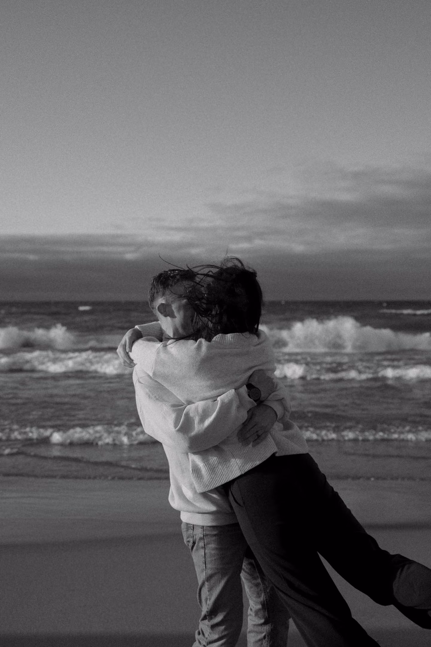 Couple embracing on a beach with waves and cloudy sky in the background in black and white.