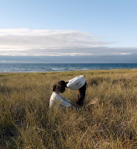 Two people in tall grass by the sea, one bending toward the other under blue sky with clouds.