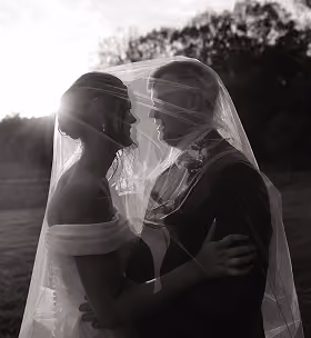Silhouetted bride and groom face each other under a veil outdoors at sunset.