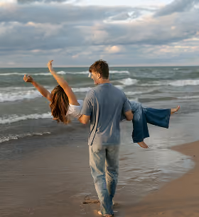 Man carrying a woman while walking barefoot on a sandy beach by the ocean under a cloudy sky.