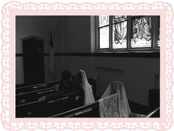 Two people sitting in church pews with lace fabric draped over them near stained glass windows.