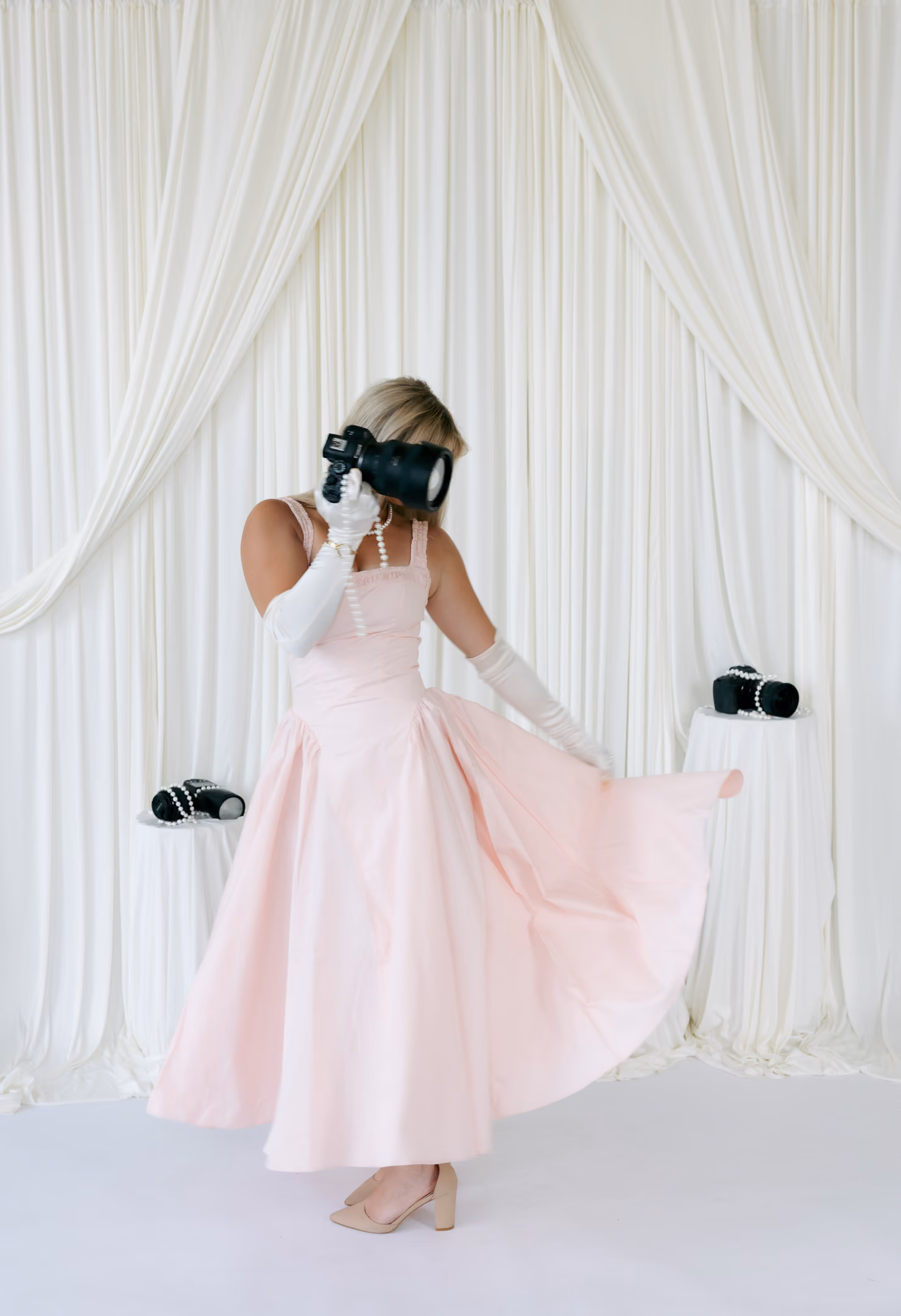 Woman in pink dress holding a camera to her face with white curtains and cameras on tables behind her.