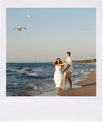 Couple holding hands and walking barefoot on a sandy beach with seagulls flying above.