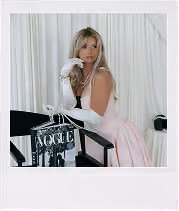 Woman in a pale pink dress with white gloves and pearl necklace sitting on a black director’s chair.