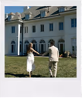 Couple in formal attire holding hands and walking on grass toward a large white mansion.