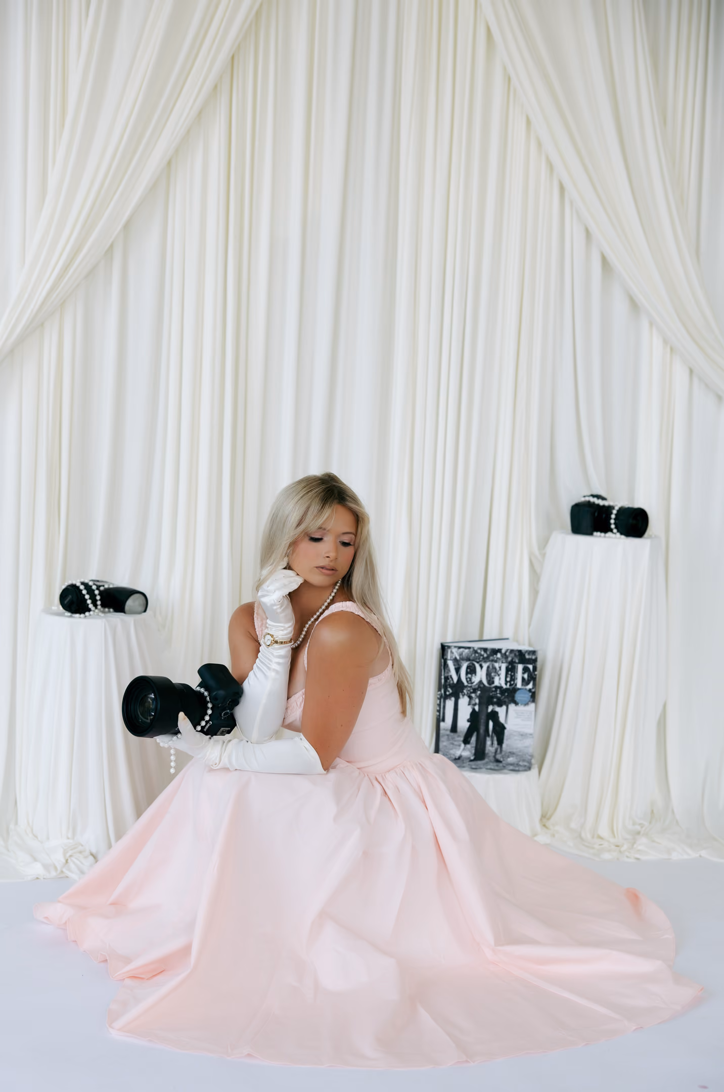 Woman in pink gown and white gloves holding a camera with pearls, seated before white curtains with Vogue magazine.