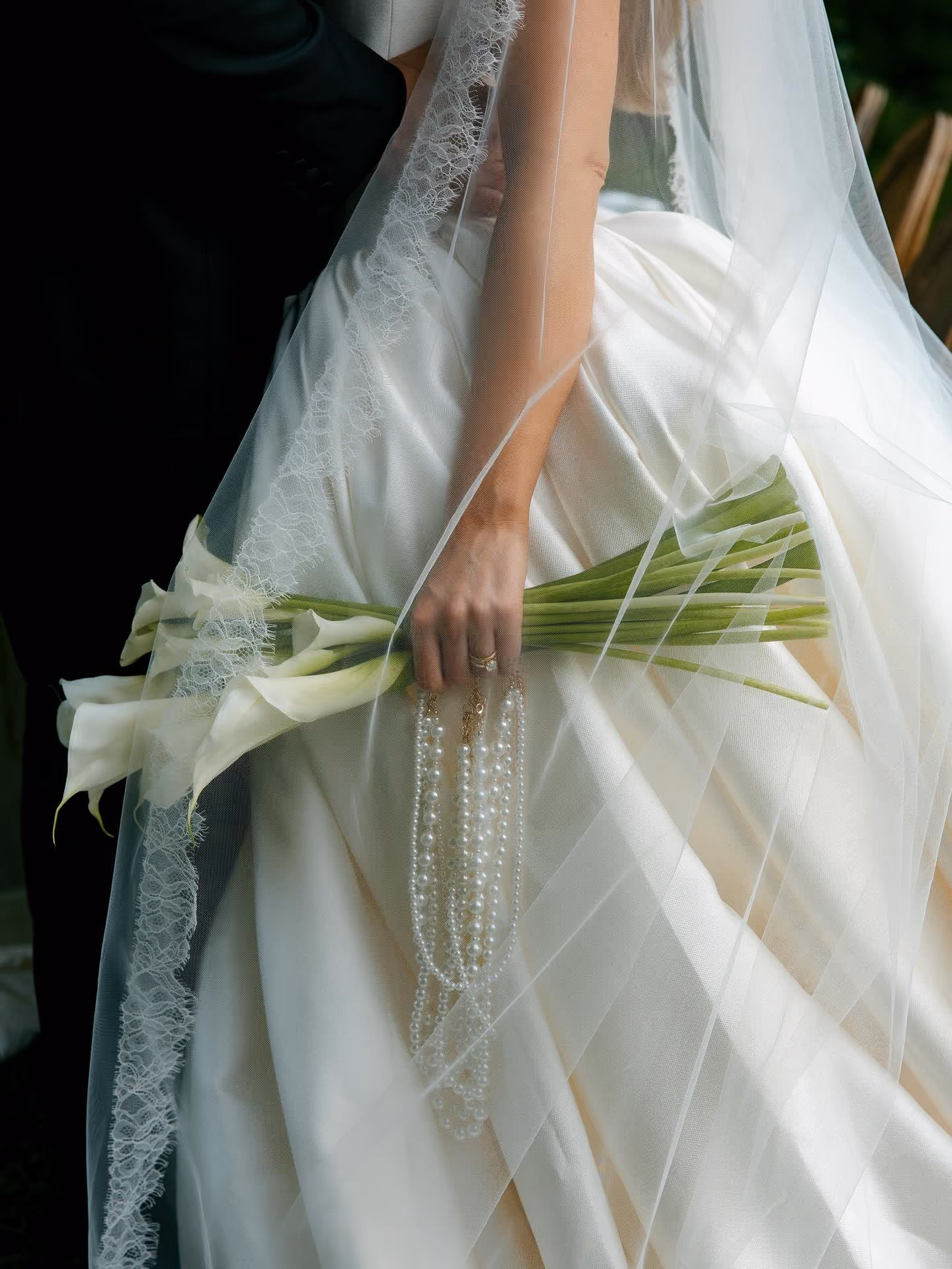 Close-up of bride in white gown holding a calla lily bouquet and pearl necklace under lace veil.